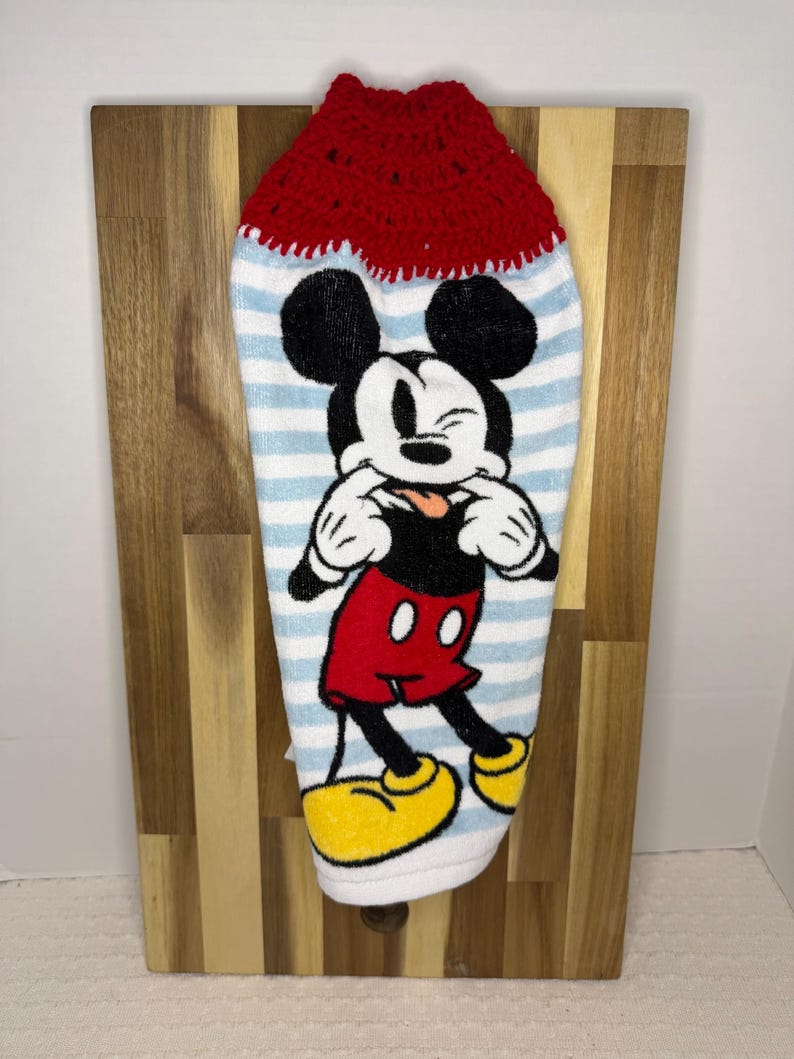May include: A kitchen towel with a red crocheted top and a blue and white striped body. The towel features a large graphic of Mickey Mouse in his classic outfit. The towel is hanging on a wooden board.