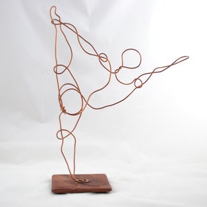 May include: A copper wire sculpture of a person in a yoga pose, standing on a wooden base.