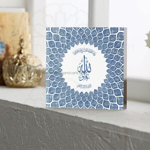 May include: A square, white canvas print with a blue and white design. The design features a circle with the Arabic word "Allah" in the center, surrounded by a pattern of blue leaves. The leaves are arranged in a circular pattern around the circle.