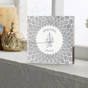May include: A silver acrylic block with a white background and a silver geometric pattern. The block features a silver Arabic calligraphy design with the word "Allah" in the center. The block is a decorative piece for a home or office.