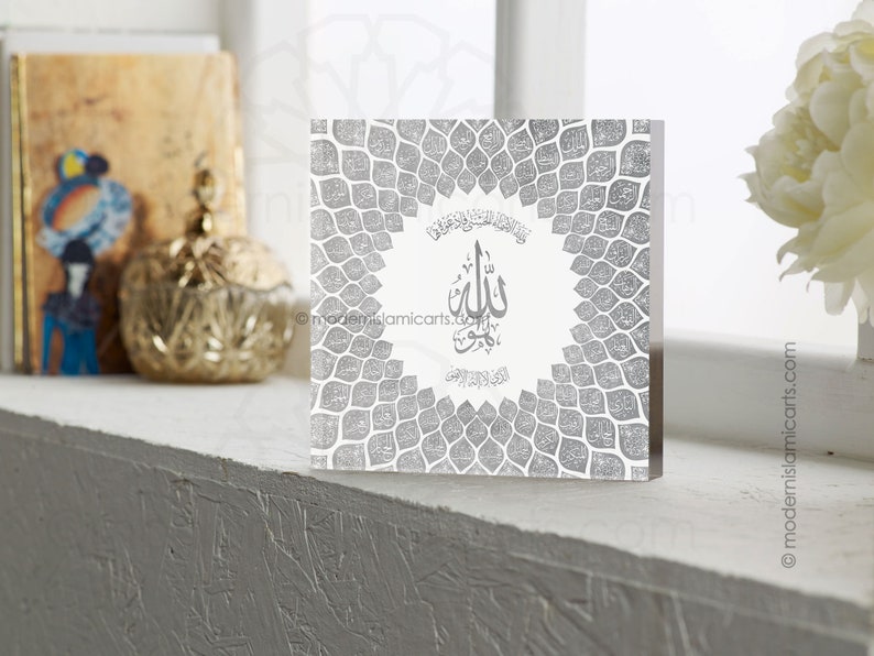 May include: A silver acrylic block with a white background and a silver geometric pattern. The block features a silver Arabic calligraphy design with the word "Allah" in the center. The block is a decorative piece for a home or office.