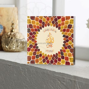 May include: A square wall art piece with a beige background and a circular design of colorful leaves. The leaves are in shades of red, orange, yellow, and brown. The center of the design features a white circle with Arabic calligraphy in black.