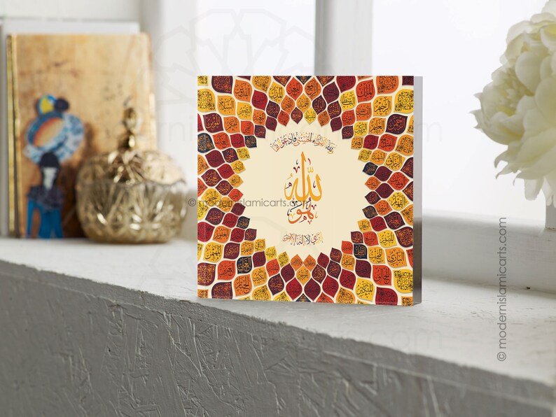 May include: A square wall art piece with a beige background and a circular design of colorful leaves. The leaves are in shades of red, orange, yellow, and brown. The center of the design features a white circle with Arabic calligraphy in black.