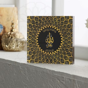 May include: A square black and gold wall art piece with an intricate design of Arabic calligraphy. The design features a circular pattern with the word "Allah" in the center. The background is black with gold accents.