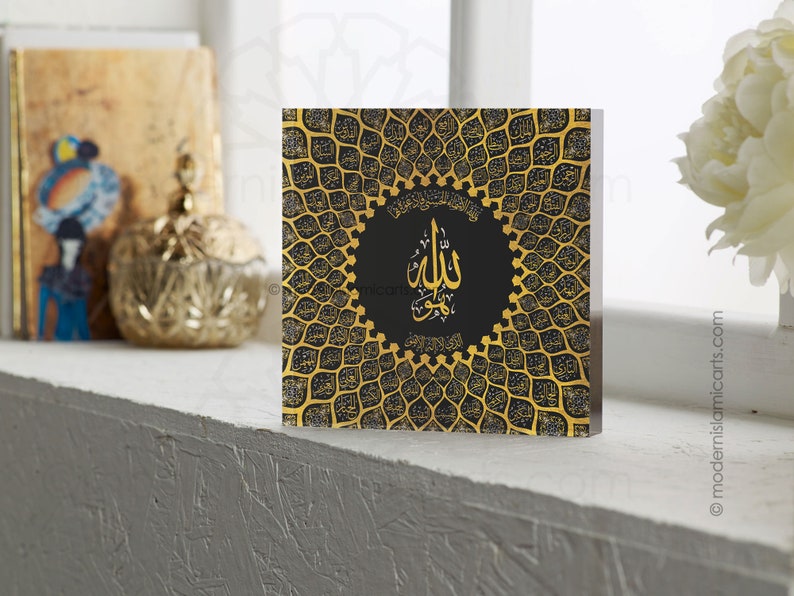 May include: A square black and gold wall art piece with an intricate design of Arabic calligraphy. The design features a circular pattern with the word "Allah" in the center. The background is black with gold accents.