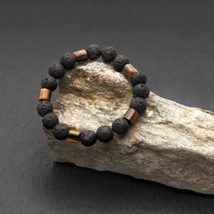 Può includere: Un braccialetto di perline di lava nera e occhio di tigre su una roccia grigia.