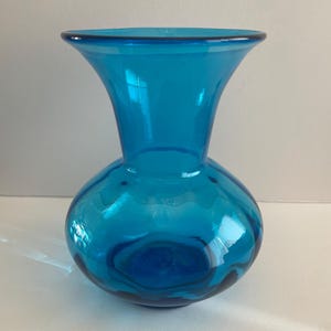 May include: A vibrant turquoise glass vase with a rounded base and flared top. The vase has a smooth, glossy finish and is designed for displaying flowers or as a decorative accent. The vase is approximately 8 inches tall.