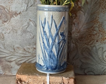 Puede incluir: Un jarrón cilíndrico de cerámica con acabado de esmalte salino en color beige claro, con patrones botánicos azules en relieve de hierbas altas y una libélula. El jarrón tiene una superficie texturizada y una banda geométrica decorativa en los bordes.