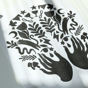May include: Black and white print of two hands with various symbols and designs surrounding them, including stars, moons, flowers, and a cat.