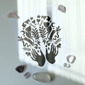 May include: Black and white print of two hands holding a variety of symbols, including stars, moons, flowers, and eyes. The print is on a white background.