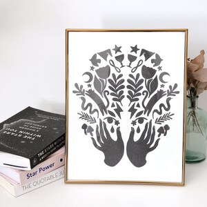 May include: A black and white framed print with a symmetrical design of two hands holding a variety of symbols, including stars, moons, flowers, and leaves.