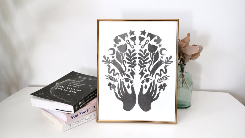 May include: A black and white framed print with a symmetrical design of two hands holding a variety of symbols, including stars, moons, flowers, and leaves.