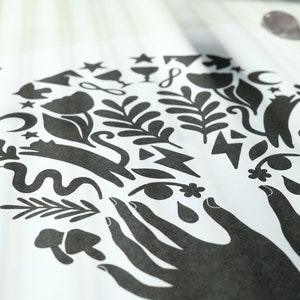 May include: Black and white print of two hands holding a variety of symbols, including flowers, stars, moons, snakes, and leaves.