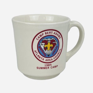 Könnte beinhalten: Weiße Keramiktasse mit einem roten, blauen und gelben Logo, auf dem "Camp Bert Adams Atlanta Area Council Diamond Jubilee 1985 Summer Camp" steht.