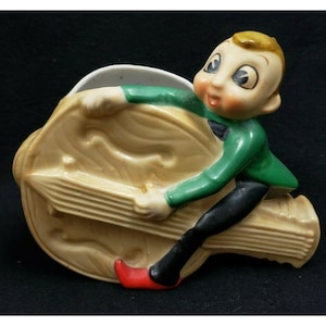 Puede incluir: Un macetero de cerámica con forma de arpa con una figura de elfo verde y negro sentada encima. El elfo está sujetando el arpa y tiene un zapato rojo.