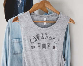 Peut inclure: Un débardeur gris avec le texte "BASEBALL MOM" en noir, avec le numéro "15" en dessous. Le débardeur est suspendu à un cintre en bois, avec une chemise en jean et un chapeau noir en arrière-plan.
