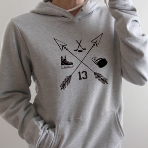 May include: A gray hoodie with a front pocket and a drawstring hood. The front features a black graphic of crossed arrows, a hockey skate, a hockey puck, a hockey stick and the number 13.