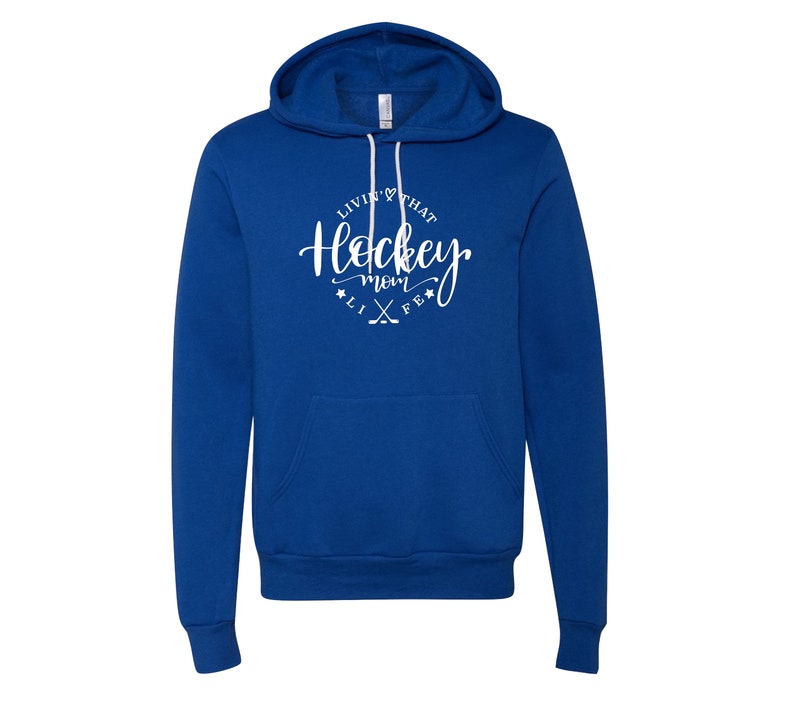 K&ouml;nnte beinhalten: Ein k&ouml;nigsblauer Hoodie mit Fronttasche und Kapuze mit Kordelzug. Der wei&szlig;e Text auf der Vorderseite lautet "Livin' that Hockey Mom Life" in Schreibschrift, mit Hockeyschl&auml;gern und Sternen.