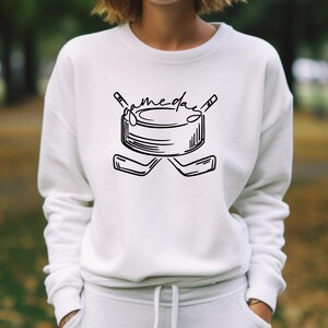 K&ouml;nnte beinhalten: Wei&szlig;es Sweatshirt mit einer schwarzen Hockey-Grafik. Das Design zeigt einen Puck, gekreuzte Hockeyschl&auml;ger und das Wort "gameday". Das Sweatshirt ist ein l&auml;ssiger Stil.