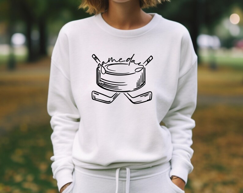 K&ouml;nnte beinhalten: Wei&szlig;es Sweatshirt mit einer schwarzen Hockey-Grafik. Das Design zeigt einen Puck, gekreuzte Hockeyschl&auml;ger und das Wort "gameday". Das Sweatshirt ist ein l&auml;ssiger Stil.