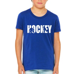 Puede incluir: Camiseta azul real con la palabra "HOCKEY" en letras blancas. La "O" en "HOCKEY" presenta siluetas de jugadores de hockey. Un joven lleva la camiseta.