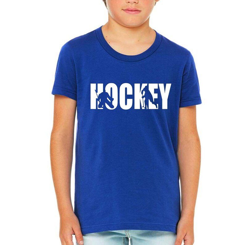 Puede incluir: Camiseta azul real con la palabra "HOCKEY" en letras blancas. La "O" en "HOCKEY" presenta siluetas de jugadores de hockey. Un joven lleva la camiseta.