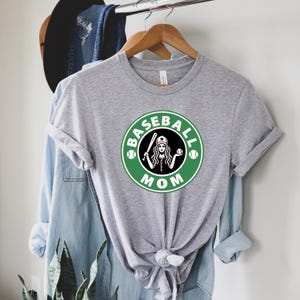 May include: A heather gray t-shirt with a green and white graphic that says "Baseball Mom." The graphic features a woman holding a baseball bat and ball inside a circle. The shirt is hanging on a wooden hanger.