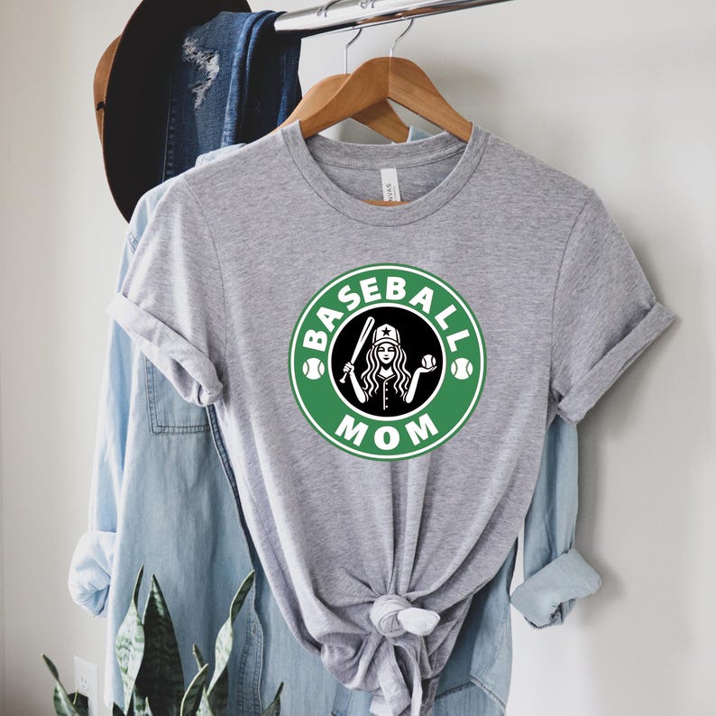 May include: A heather gray t-shirt with a green and white graphic that says "Baseball Mom." The graphic features a woman holding a baseball bat and ball inside a circle. The shirt is hanging on a wooden hanger.