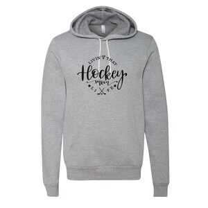 K&ouml;nnte beinhalten: Ein hellgrauer Kapuzenpullover mit Kordelzug und Vordertasche. Der Hoodie zeigt den Text "Livin' That Hockey Mom Life" in schwarzer Schrift in einem kreisf&ouml;rmigen Design mit Hockeyschl&auml;gern und Sternen.