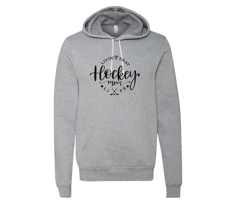 K&ouml;nnte beinhalten: Ein hellgrauer Kapuzenpullover mit Kordelzug und Vordertasche. Der Hoodie zeigt den Text "Livin' That Hockey Mom Life" in schwarzer Schrift in einem kreisf&ouml;rmigen Design mit Hockeyschl&auml;gern und Sternen.