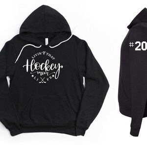 K&ouml;nnte beinhalten: Schwarzer Kapuzenpullover mit Kordelzug und Vordertasche. Auf der Vorderseite steht in wei&szlig;er Schrift "Livin' That Hockey Mom Life". Auf dem rechten &Auml;rmel ist die wei&szlig;e Nummer "20" zu sehen. Ein l&auml;ssiges Kleidungsst&uuml;ck.