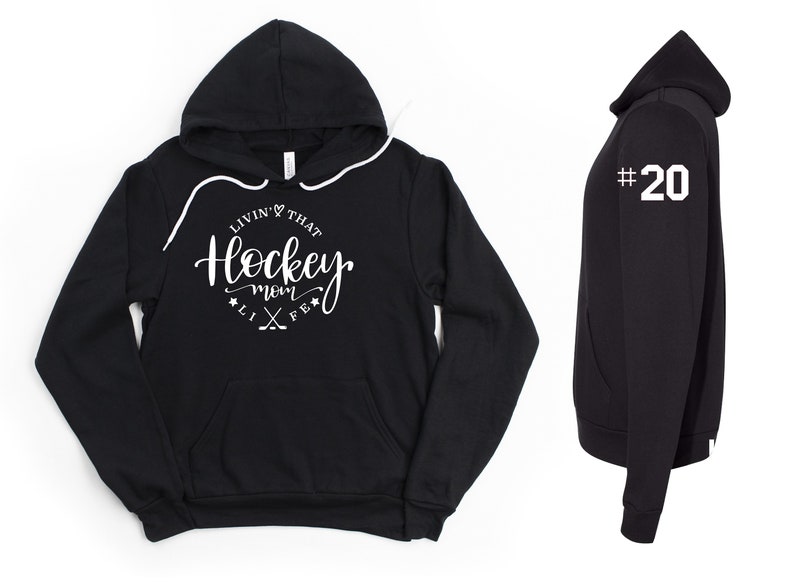 K&ouml;nnte beinhalten: Schwarzer Kapuzenpullover mit Kordelzug und Vordertasche. Auf der Vorderseite steht in wei&szlig;er Schrift "Livin' That Hockey Mom Life". Auf dem rechten &Auml;rmel ist die wei&szlig;e Nummer "20" zu sehen. Ein l&auml;ssiges Kleidungsst&uuml;ck.