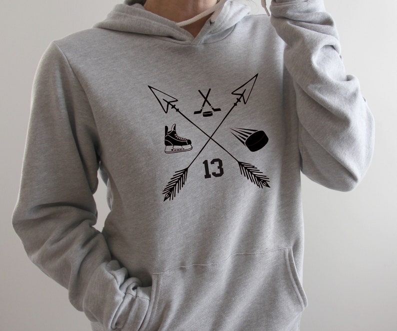 May include: A gray hoodie with a front pocket and a drawstring hood. The front features a black graphic of crossed arrows, a hockey skate, a hockey puck, a hockey stick and the number 13.