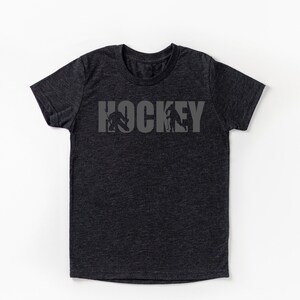 Puede incluir: Camiseta gris oscuro con la palabra "HOCKEY" en letras grises. Las letras incorporan siluetas de jugadores de hockey en acci&oacute;n. La camiseta es de cuello redondo y manga corta.
