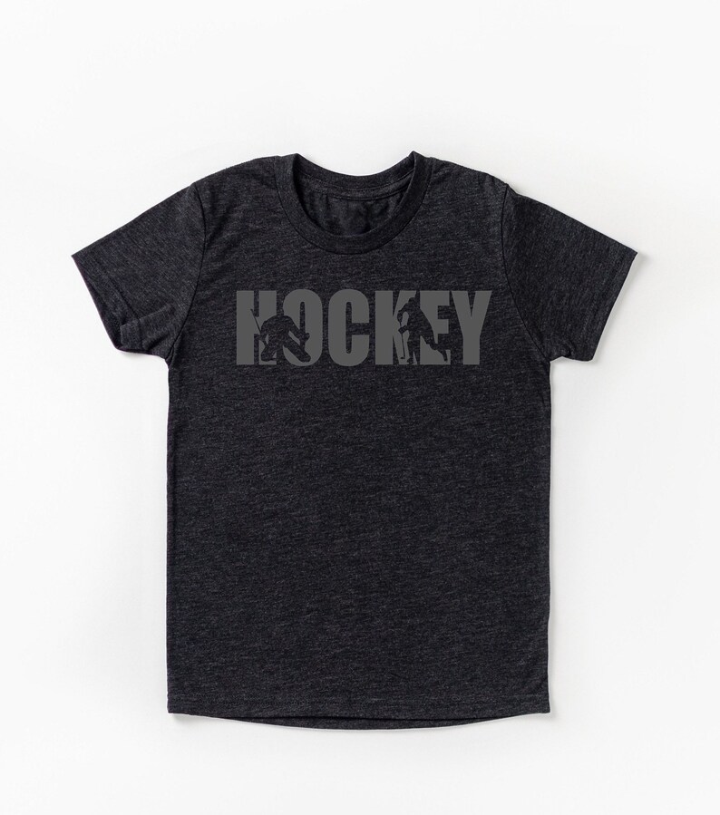 Puede incluir: Camiseta gris oscuro con la palabra "HOCKEY" en letras grises. Las letras incorporan siluetas de jugadores de hockey en acci&oacute;n. La camiseta es de cuello redondo y manga corta.
