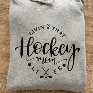 K&ouml;nnte beinhalten: Ein gefalteter hellgrauer Kapuzenpullover mit wei&szlig;em Kordelzug. Auf der Vorderseite des Hoodies steht in schwarzer Schrift und Blockbuchstaben der Schriftzug "Livin' That Hockey Mom Life", mit Hockeyschl&auml;gern und Sternen.