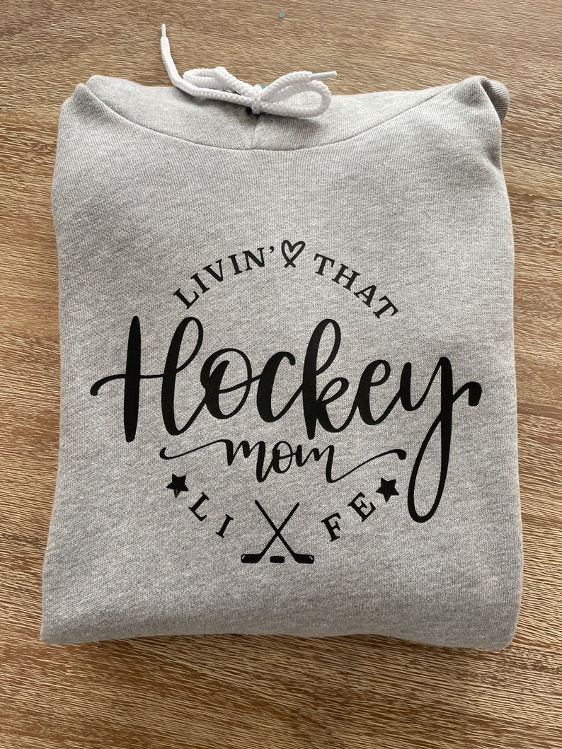 K&ouml;nnte beinhalten: Ein gefalteter hellgrauer Kapuzenpullover mit wei&szlig;em Kordelzug. Auf der Vorderseite des Hoodies steht in schwarzer Schrift und Blockbuchstaben der Schriftzug "Livin' That Hockey Mom Life", mit Hockeyschl&auml;gern und Sternen.