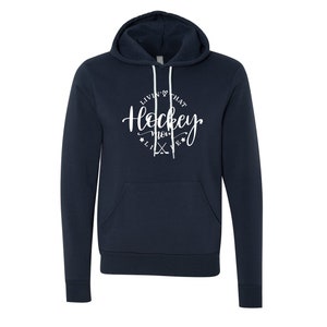 K&ouml;nnte beinhalten: Marineblauer Kapuzenpullover mit wei&szlig;em Kordelzug und Vordertasche. Der Kapuzenpullover zeigt den Text "Livin' that Hockey Mom Life" in einem kreisf&ouml;rmigen Design mit Hockeyschl&auml;gern und Sternen. Ein bequemes, l&auml;ssiges Oberteil.