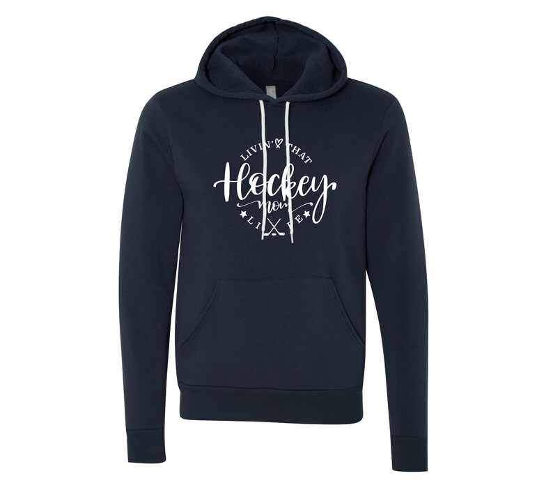K&ouml;nnte beinhalten: Marineblauer Kapuzenpullover mit wei&szlig;em Kordelzug und Vordertasche. Der Kapuzenpullover zeigt den Text "Livin' that Hockey Mom Life" in einem kreisf&ouml;rmigen Design mit Hockeyschl&auml;gern und Sternen. Ein bequemes, l&auml;ssiges Oberteil.