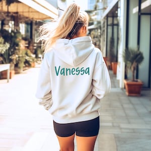 Puede incluir: Una sudadera con capucha color crema con el nombre "Vanessa" impreso en verde azulado en la espalda. La sudadera tiene una capucha con cord&oacute;n y mangas largas. La persona lleva pantalones cortos negros.