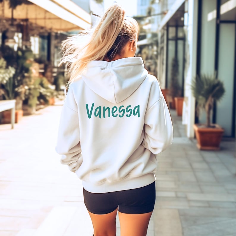 Puede incluir: Una sudadera con capucha color crema con el nombre "Vanessa" impreso en verde azulado en la espalda. La sudadera tiene una capucha con cord&oacute;n y mangas largas. La persona lleva pantalones cortos negros.