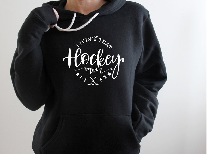 K&ouml;nnte beinhalten: Schwarzer Kapuzenpullover mit wei&szlig;em Kordelzug und Vordertasche. Der Kapuzenpullover hat eine wei&szlig;e Grafik mit der Aufschrift "Livin' That Hockey Mom Life" in einem kreisf&ouml;rmigen Design mit Hockeyschl&auml;gern und Sternen.