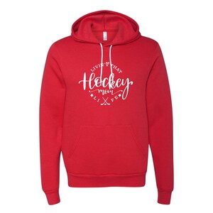 K&ouml;nnte beinhalten: Ein roter Kapuzenpullover mit wei&szlig;em Kordelzug und Vordertasche. Der Kapuzenpullover hat eine wei&szlig;e kreisf&ouml;rmige Grafik mit dem Text "Livin' that Hockey Mom Life" und gekreuzten Hockeyschl&auml;gern.