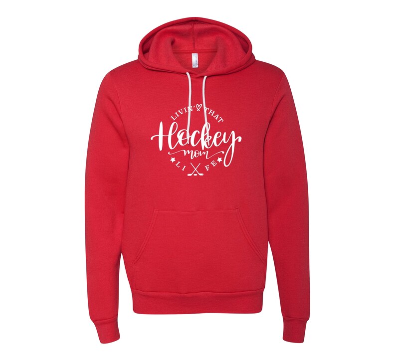 K&ouml;nnte beinhalten: Ein roter Kapuzenpullover mit wei&szlig;em Kordelzug und Vordertasche. Der Kapuzenpullover hat eine wei&szlig;e kreisf&ouml;rmige Grafik mit dem Text "Livin' that Hockey Mom Life" und gekreuzten Hockeyschl&auml;gern.