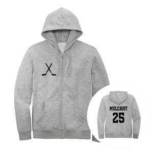 May include: A light gray zip-up hoodie featuring a black graphic of two crossed hockey sticks on the left chest. The back view, shown in a circular inset, displays the name MULCAHY and the number 25 in bold black lettering on the back.
