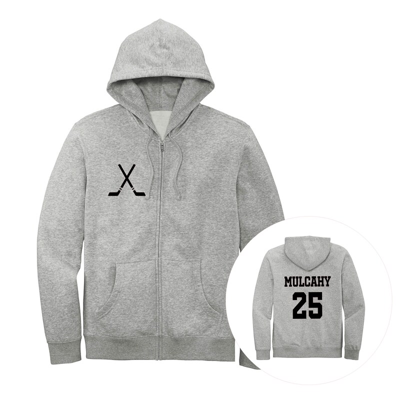 May include: A light gray zip-up hoodie featuring a black graphic of two crossed hockey sticks on the left chest. The back view, shown in a circular inset, displays the name MULCAHY and the number 25 in bold black lettering on the back.