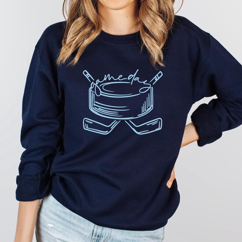K&ouml;nnte beinhalten: Marineblaues Sweatshirt mit hellblauer Hockey-Grafik. Das Design zeigt einen Hockeypuck, gekreuzte Hockeyschl&auml;ger und den Text "gameday". Ein l&auml;ssiges Oberteil f&uuml;r Sportfans.