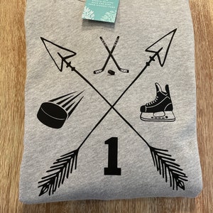 May include: A gray hoodie with a black hockey-themed design. The design includes crossed arrows, hockey sticks, a puck, a skate, and the number 1. A tag with the text "Our Wildflowers" is attached.