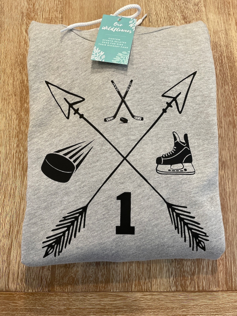 May include: A gray hoodie with a black hockey-themed design. The design includes crossed arrows, hockey sticks, a puck, a skate, and the number 1. A tag with the text "Our Wildflowers" is attached.