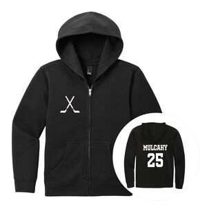 May include: A black zip-up hoodie featuring a white graphic of two crossed hockey sticks on the left chest. An inset circle shows the back of the hoodie, which displays the name MULCAHY in white block letters above the number 25 in white.